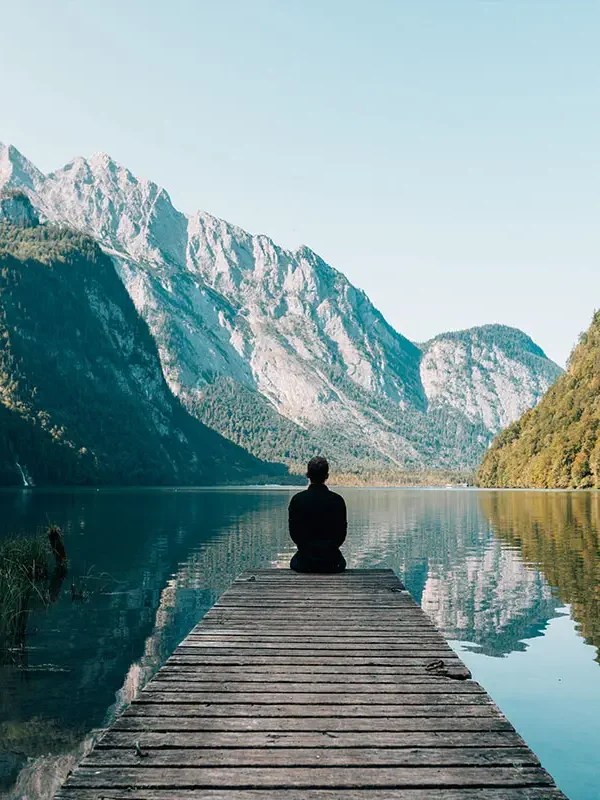 Person sitting on a dock facing a serene mountain lake scene.