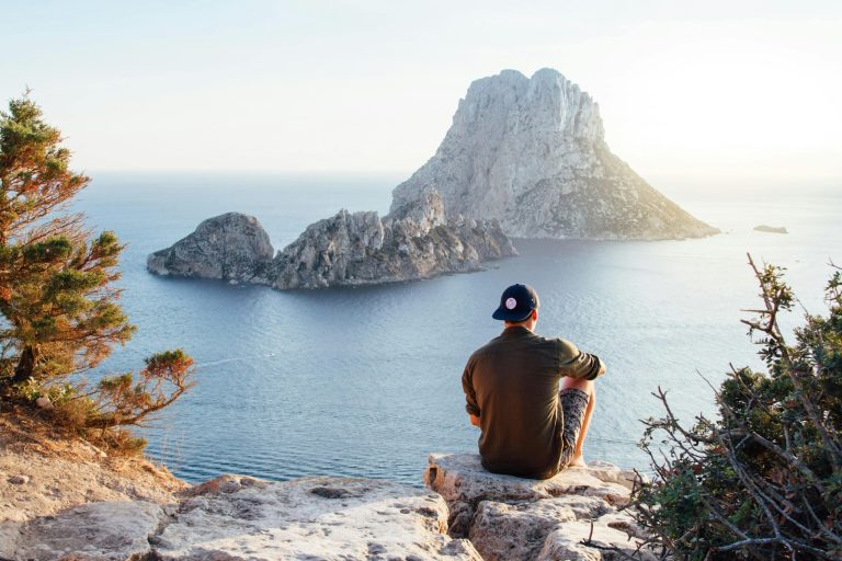 Person sitting on cliff overlooking ocean and large rock formation.