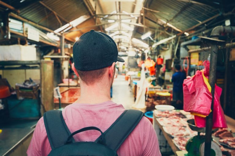 Man with backpack walks through busy indoor market with meat stalls.