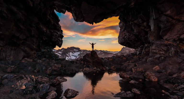 Person standing in cave opening with colorful sunset and mountains in background.