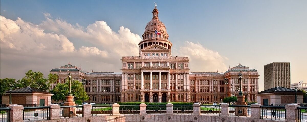 State capitol building with a large dome and surrounding gardens under a partly cloudy sky.