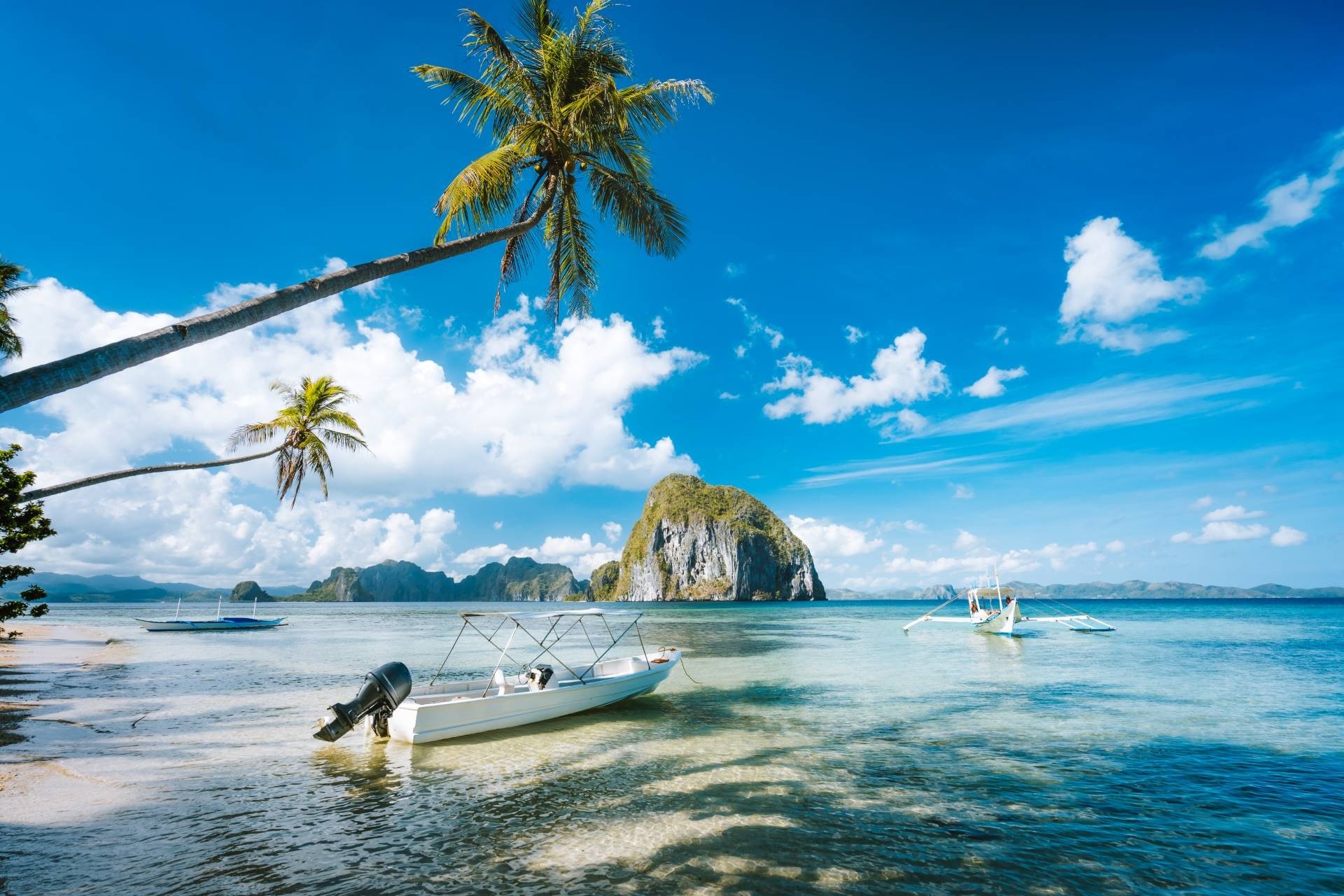 Tropical beach with boats, palm trees, and a limestone island under a blue sky.