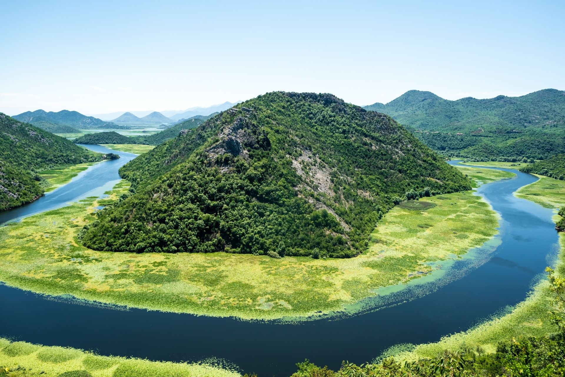 River bends around a lush, green hill with mountains in the background.