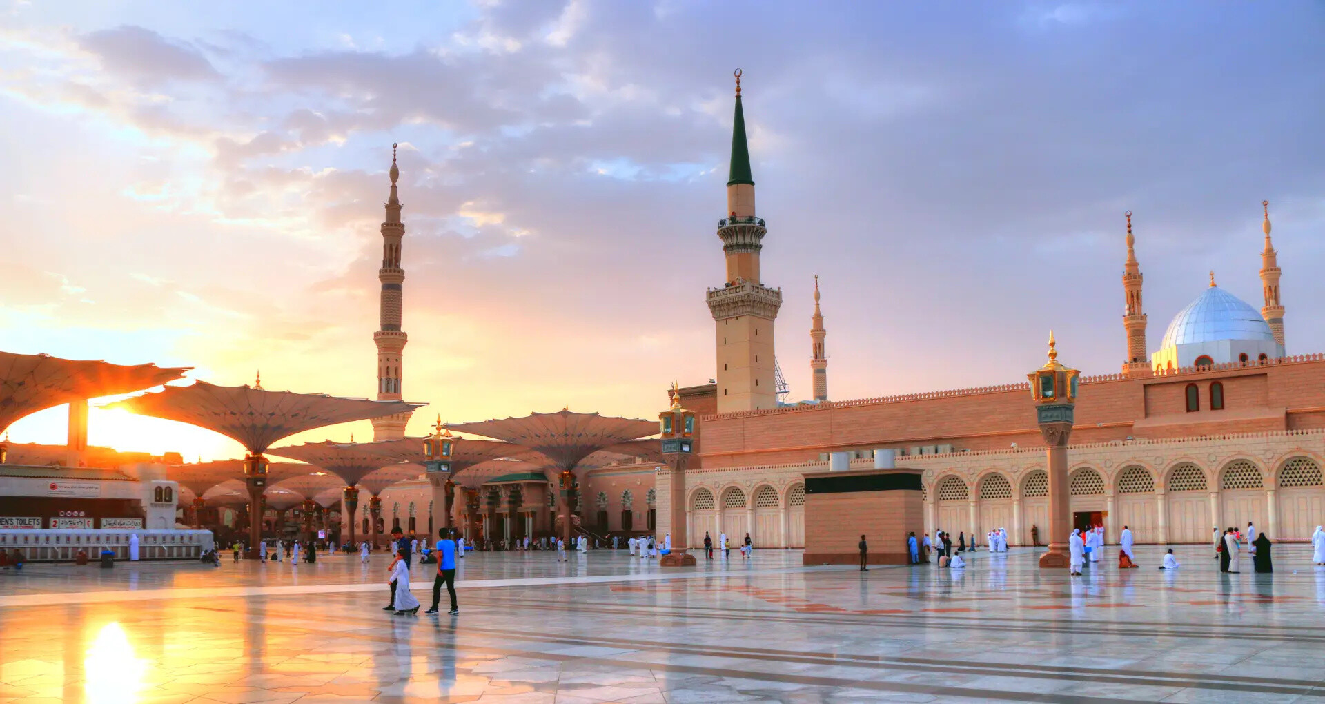 People at a mosque courtyard with tall minarets and a sunset sky.